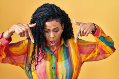 Orta yaşlı İspanyol bir kadın sarı arka planda dikiliyor parmaklarını göstererek reklamını yapıyor, şaşırmış bir yüz ve ağzı açık. 