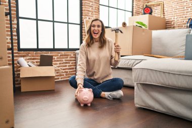 Young woman holding piggy bank and hammer moving to a new home celebrating crazy and amazed for success with open eyes screaming excited. 