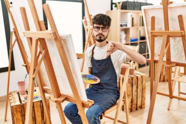 Hispanic man with beard at art studio with angry face, negative sign showing dislike with thumbs down, rejection concept 