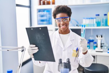 African american woman wearing scientist uniform reading document holding test tubes at laboratory