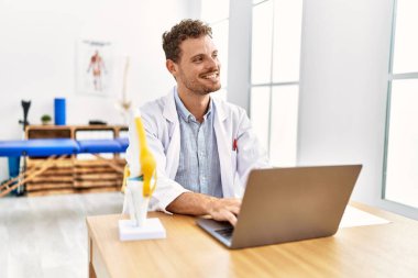Klinikte çalışan dizüstü bilgisayar kullanan fizyoterapist üniformalı genç bir İspanyol.