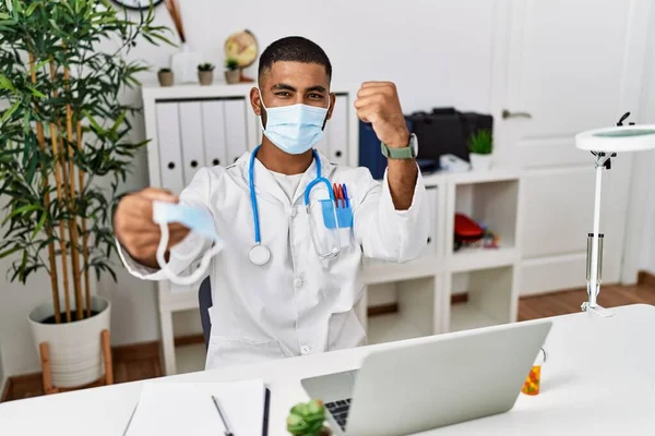 Young indian doctor offering safety mask annoyed and frustrated shouting with anger, yelling crazy with anger and hand raised