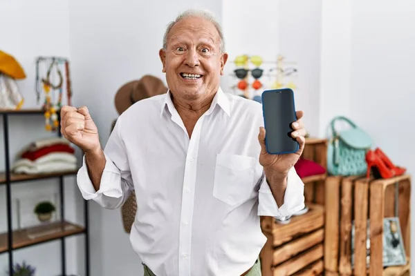 Senior man holding smartphone at retail shop screaming proud, celebrating victory and success very excited with raised arms