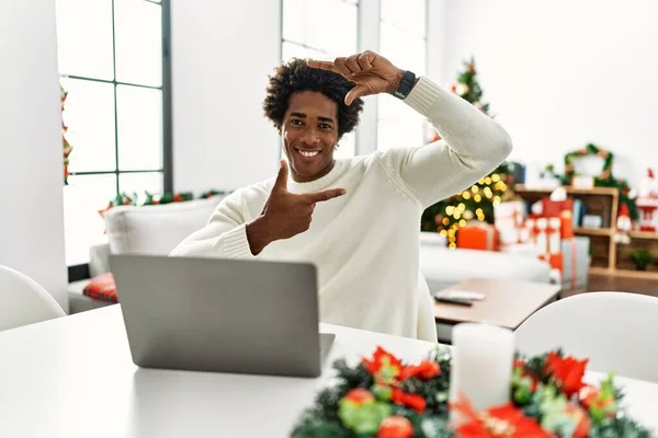 クリスマスツリーの笑顔でテーブルの上に座ってノートパソコンを使用して若いアフリカ系アメリカ人の男性は幸せな顔を持つ手や指でフレームを作ります 創造性と写真の概念 — ストック写真