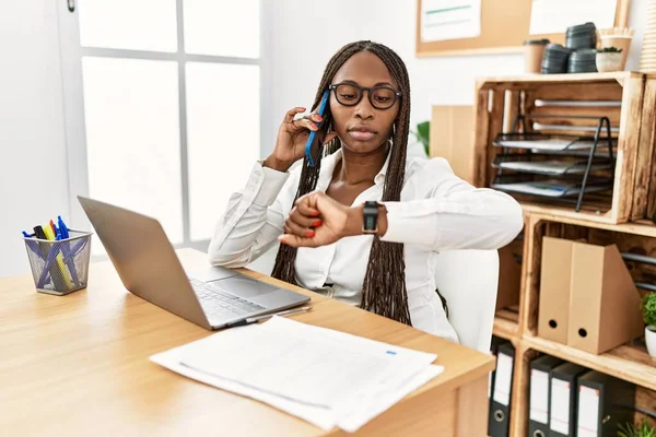 Black woman with braids working at the office speaking on the phone checking the time on wrist watch, relaxed and confident