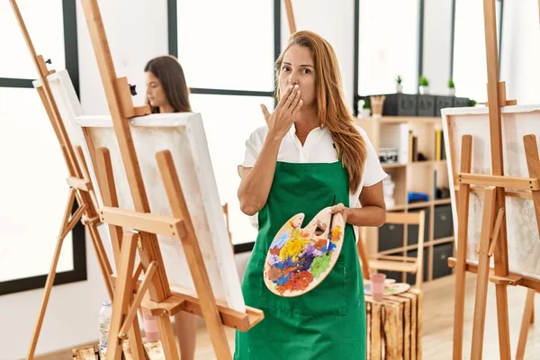 Young hispanic middle age woman at art classroom covering mouth with hand, shocked and afraid for mistake. surprised expression