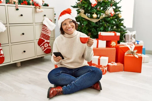 中年ヒスパニック女性使用スマートフォンと飲むコーヒーによって座ってクリスマスツリーに家 — ストック写真