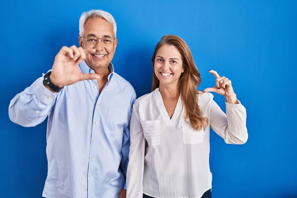 Middle age hispanic couple standing over blue background smiling and confident gesturing with hand doing small size sign with fingers looking and the camera. measure concept.