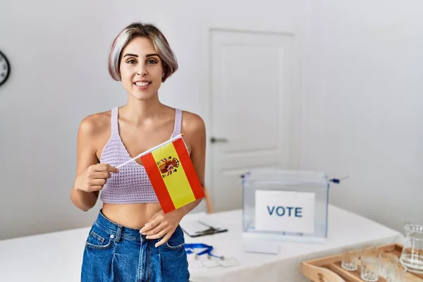 Young beautiful woman at political campaign election holding spain flag looking positive and happy standing and smiling with a confident smile showing teeth