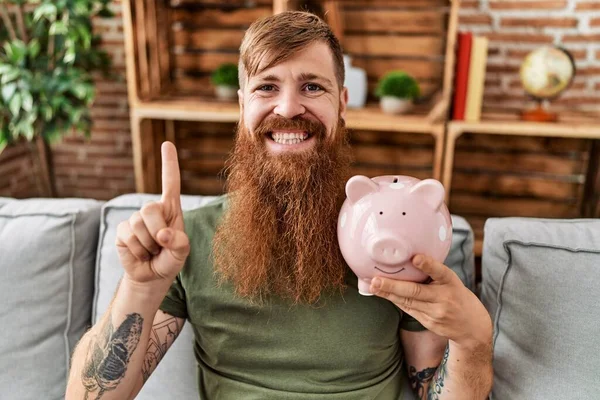 Redhead Man Holding Piggy Bank Smiling Idea Question Pointing Finger — Fotografia de Stock