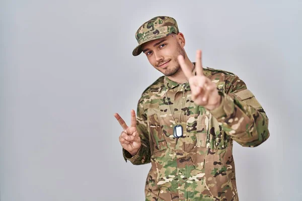 Young hispanic man wearing camouflage army uniform smiling looking to the camera showing fingers doing victory sign. number two.