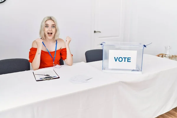 Young caucasian woman at political election sitting by ballot screaming proud, celebrating victory and success very excited with raised arms