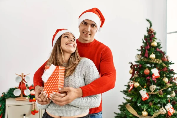 Молодая Пара Обнимается Улыбается Счастливой Ношение Рождественской Шляпы Подарком Дома — стоковое фото