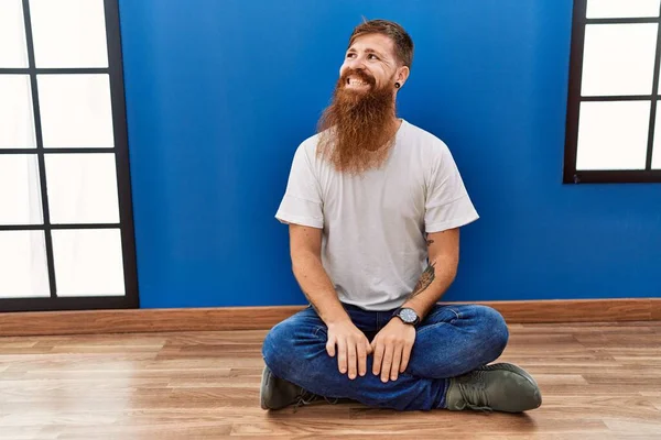 Redhead man with long beard sitting on the floor at empty room looking away to side with smile on face, natural expression. laughing confident.