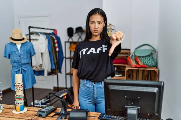 Young hispanic woman working as staff at retail boutique looking unhappy and angry showing rejection and negative with thumbs down gesture. bad expression.