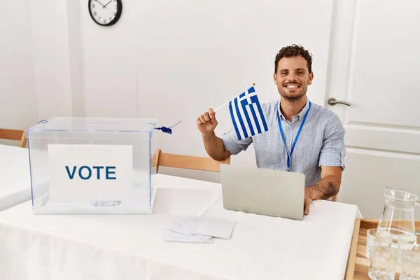 Joven Hispano Sonriendo Confiado Sosteniendo Bandera Griega Trabajando Colegio Electoral —  Fotos de Stock