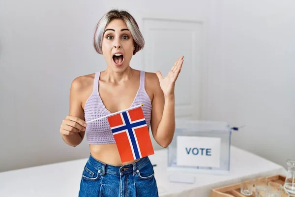 Young beautiful woman at political campaign election holding norway flag celebrating victory with happy smile and winner expression with raised hands