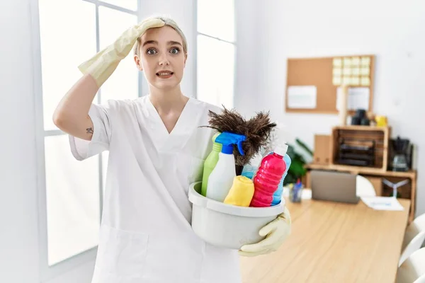 Young caucasian woman wearing cleaner uniform holding cleaning products cleaning office stressed and frustrated with hand on head, surprised and angry face