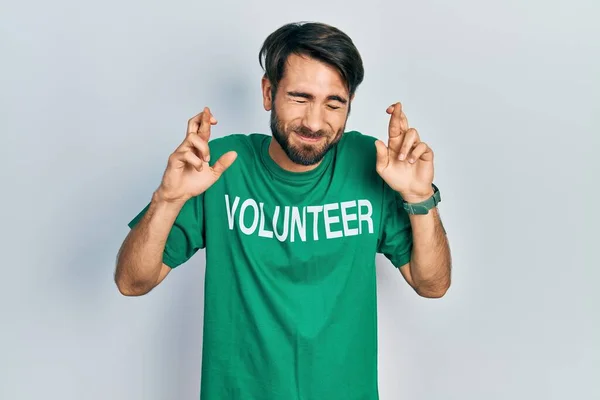 Jovem Hispânico Vestindo Voluntário Shirt Gesto Dedo Cruzado Sorrindo Com — Fotografia de Stock