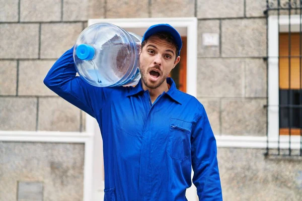 Young hispanic man holding a gallon bottle of water for delivery scared and amazed with open mouth for surprise, disbelief face