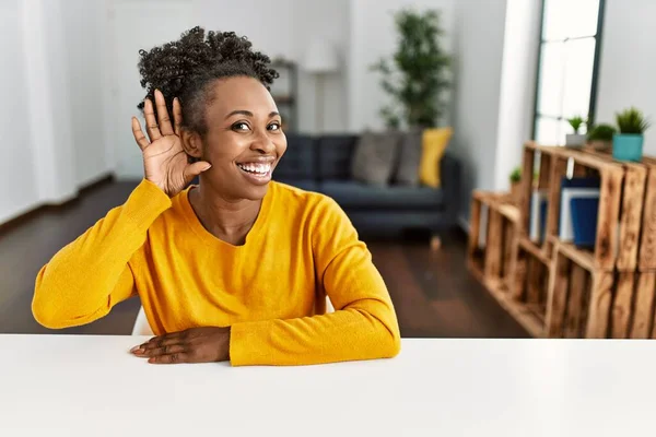 Young african american woman wearing casual clothes sitting on the table at home smiling with hand over ear listening an hearing to rumor or gossip. deafness concept.