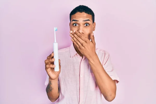 Young african american guy holding electric toothbrush covering mouth with hand, shocked and afraid for mistake. surprised expression