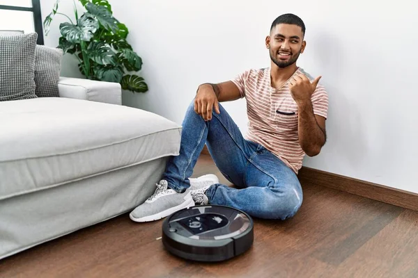 Young indian man sitting at home by vacuum robot smiling with happy face looking and pointing to the side with thumb up.