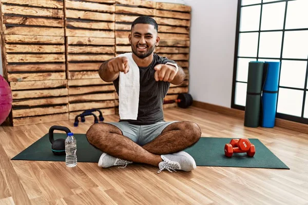 Young indian man sitting on training mat at the gym pointing to you and the camera with fingers, smiling positive and cheerful