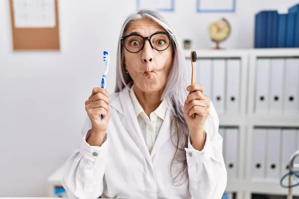 Middle age grey-haired woman working at dentist clinic holding electric and recycled teethbrush making fish face with mouth and squinting eyes, crazy and comical.