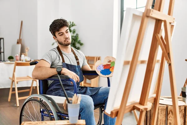 Young hispanic man sitting on wheelchair painting at art studio looking sleepy and tired, exhausted for fatigue and hangover, lazy eyes in the morning.