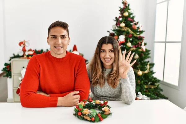 Young hispanic couple sitting at the table on christmas showing and pointing up with fingers number four while smiling confident and happy.