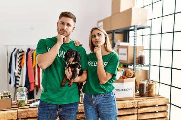 Young couple with cute dog wearing volunteer t shirt at donations stand serious face thinking about question with hand on chin, thoughtful about confusing idea
