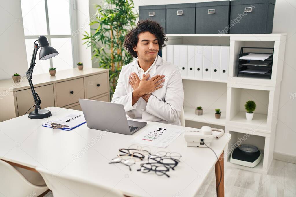 Hispanic man with curly hair working at optician office smiling with hands on chest, eyes closed with grateful gesture on face. health concept. 