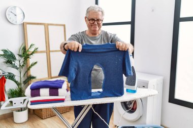 Senior caucasian man ironing holding burned iron shirt at laundry room smiling with a happy and cool smile on face. showing teeth. 