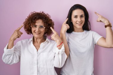 Hispanic mother and daughter wearing casual white t shirt over pink background smiling pointing to head with both hands finger, great idea or thought, good memory 