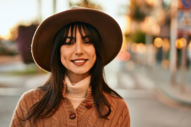 Kışlık şapkalı esmer kadın gün batımında şehirde gülümsüyor.