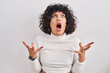 Hispanic woman with curly hair standing over isolated background crazy and mad shouting and yelling with aggressive expression and arms raised. frustration concept. 