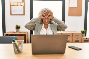 Orta yaşlı bir iş kadını ofiste dizüstü bilgisayar kullanarak çalışıyor. Dürbün gibi jest yapıyor, gözleri parmaklarından bakıyor. çılgın ifade. 