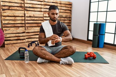 Young indian man sitting on training mat at the gym checking the time on wrist watch, relaxed and confident 