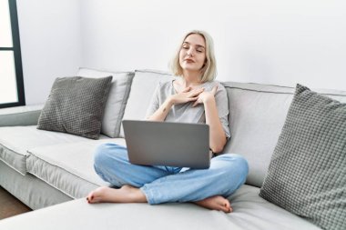 Young caucasian woman using laptop at home sitting on the sofa smiling with hands on chest with closed eyes and grateful gesture on face. health concept. 