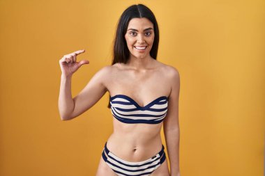 Sarı arka planda bikini giyen genç esmer kadın gülümsüyor ve el hareketleriyle küçük beden işareti yaparak kameraya bakıyor. ölçüm kavramı. 