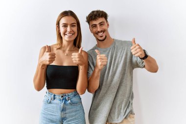 Young beautiful couple standing together over isolated background success sign doing positive gesture with hand, thumbs up smiling and happy. cheerful expression and winner gesture. 