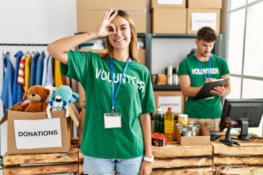 Young blonde girl wearing volunteer t shirt at donation stand doing ok gesture with hand smiling, eye looking through fingers with happy face. 