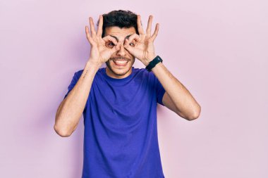 Latin asıllı genç bir adam sıradan bir tişört giyiyor dürbün gibi jest yapıyor, gözleri parmaklarından bakıyor. çılgın ifade. 