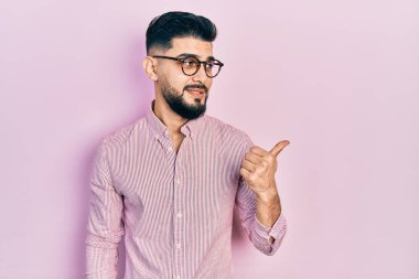 Handsome man with beard wearing casual shirt and glasses smiling with happy face looking and pointing to the side with thumb up. 