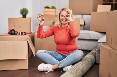 Blonde woman holding keys of new home sitting on the floor smiling happy doing ok sign with hand on eye looking through fingers 