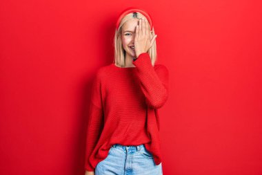Güzel sarışın bir kadın, bir gözünü elinle kapayarak gündelik kırmızı bir kazak giyiyor, yüzünde kendine güvenen bir gülümseme ve sürpriz bir duygu var.. 