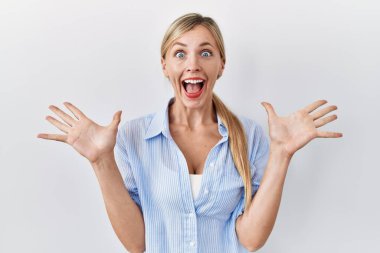 Beautiful blonde woman standing over white background celebrating crazy and amazed for success with arms raised and open eyes screaming excited. winner concept 