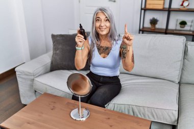 Middle age grey-haired woman using night serum sitting on the sofa smiling with an idea or question pointing finger with happy face, number one 