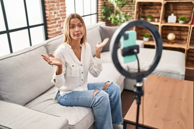 Young blonde woman recording vlog tutorial with smartphone at home shouting and screaming loud to side with hand on mouth. communication concept. 